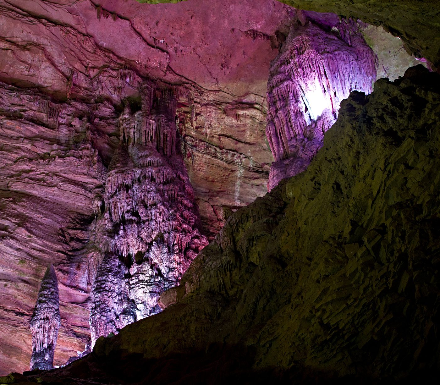 Interior of Huanglong Cave near Zhangjiajie: a tall flowstone column and hanging stalactites lit in pink and violet — Photo: Dimain69 / CC BY-SA 3.0 via Wikimedia Commons