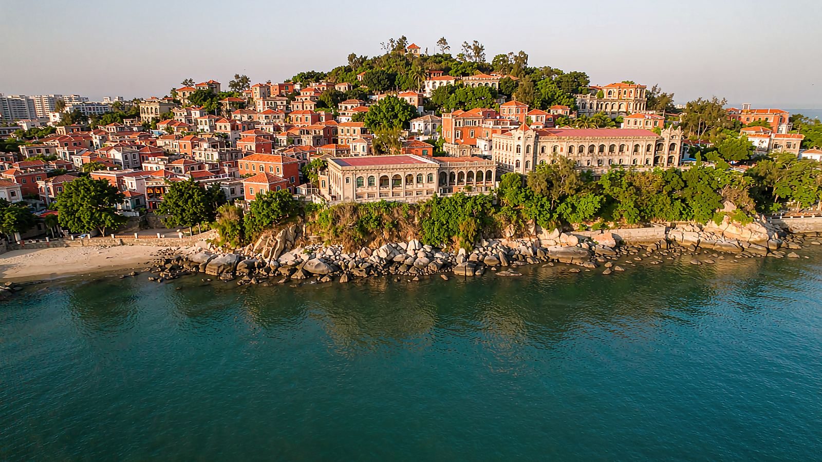 Aerial view of Gulangyu island in Xiamen: red-tiled rooftops and colonial villas on a wooded hill above a turquoise bay